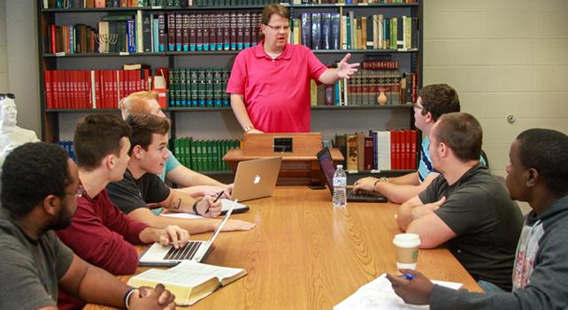 A man in a red shirt is speaking at a lectern in front of a table with seven seated individuals. Bookshelves filled with books are in the background.